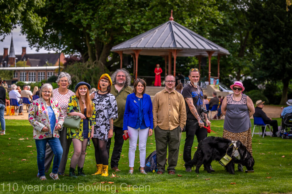 9 people stand in a group in a green park, in front of a bandstand, smiling at the camera
