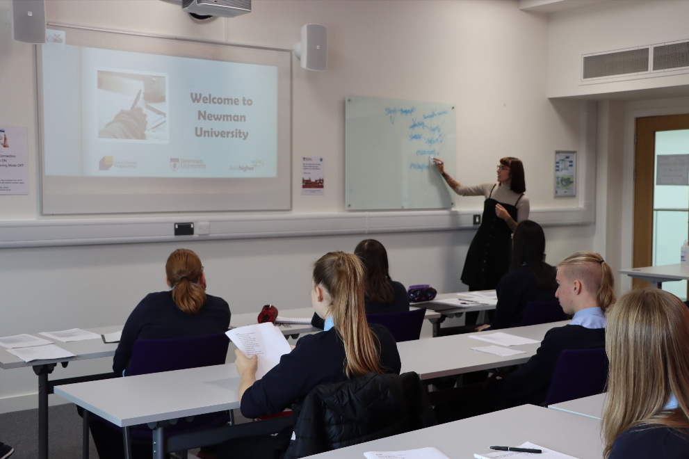Scarlett writes on a white board for a room of students - on the projector reads the words "welcome to Newman University